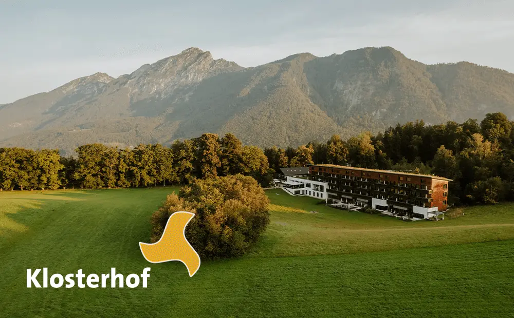 Das Bild zeigt eine Landschaft mit einem großen, grünen Feld und einem Gebäude im Vordergrund, das möglicherweise ein Hotel oder eine Unterkunft ist. Im Hintergrund erheben sich beeindruckende Berge. Das Logo „Klosterhof“ ist in der linken unteren Ecke platziert.