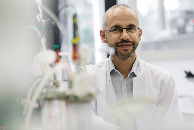 Ein lächelnder Wissenschaftler in einem Labor, gekleidet in einen weißen Laborkittel und eine Brille. Er sitzt lässig vor einer Reihe von Laborausrüstungen und schaut direkt in die Kamera. Die Atmosphäre wirkt konzentriert und professionell, mit hellen Farben im Hintergrund.