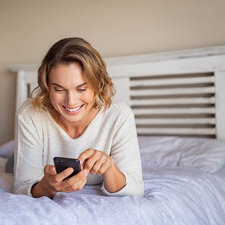 Frau liegt lächelnd mit Handy auf dem Bett
